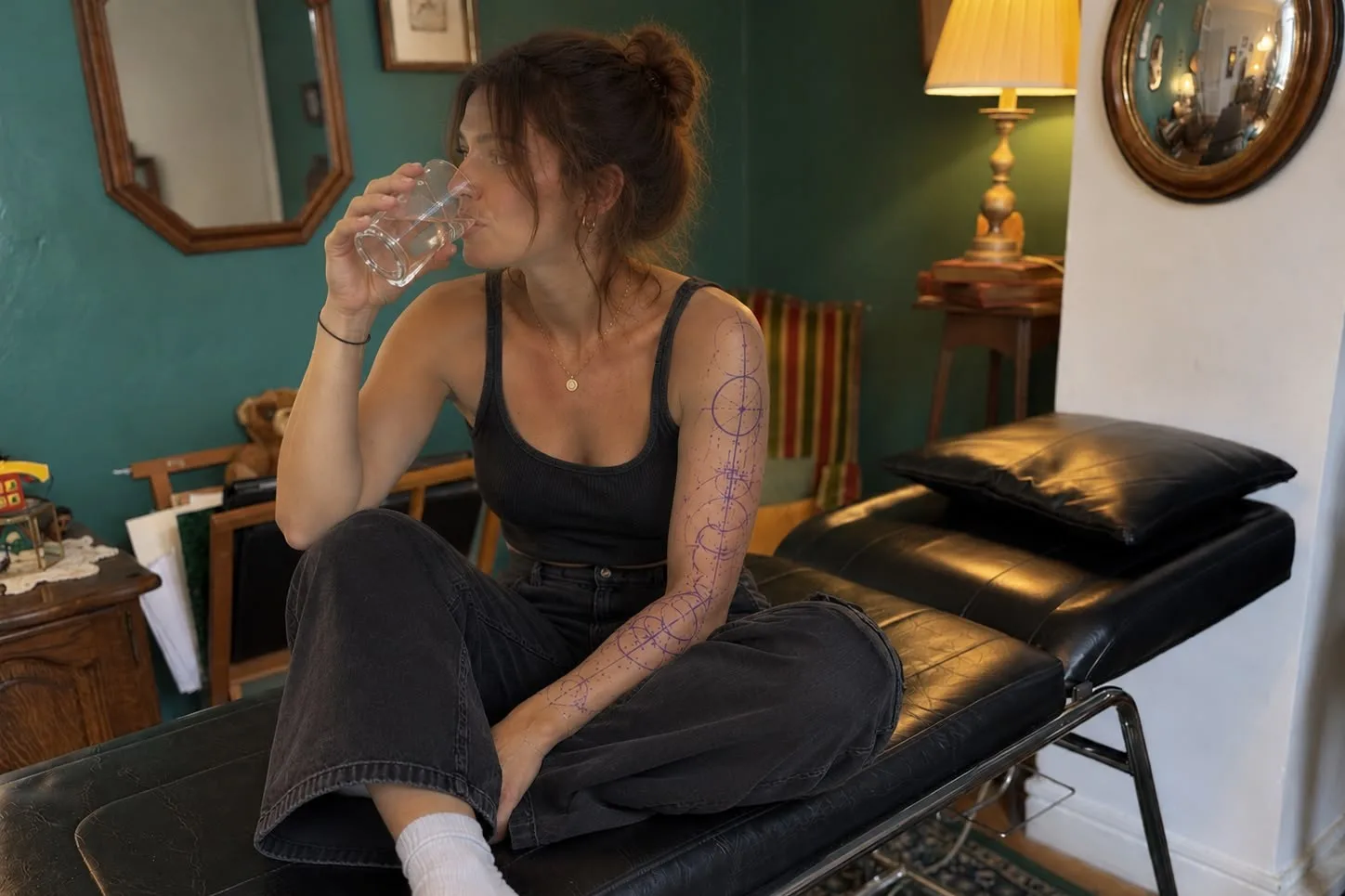 Une cliente fait une pause et boit un verre d'eau pendant sa séance, le stencil de son tatouage sur le bras, au cabinet de Barbe Noire Tattoo
